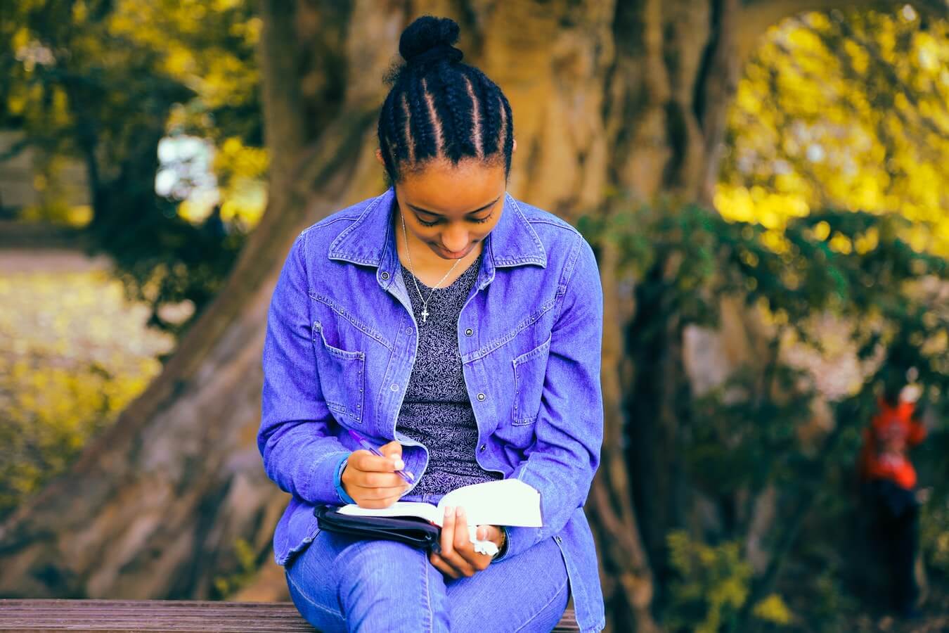 Na imagem uma menina estudando com um livro e uma caneta nas mãos